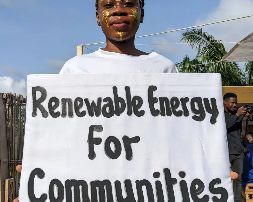 A woman is facing the camera, holding a sign saying 'renewable energy for communities'