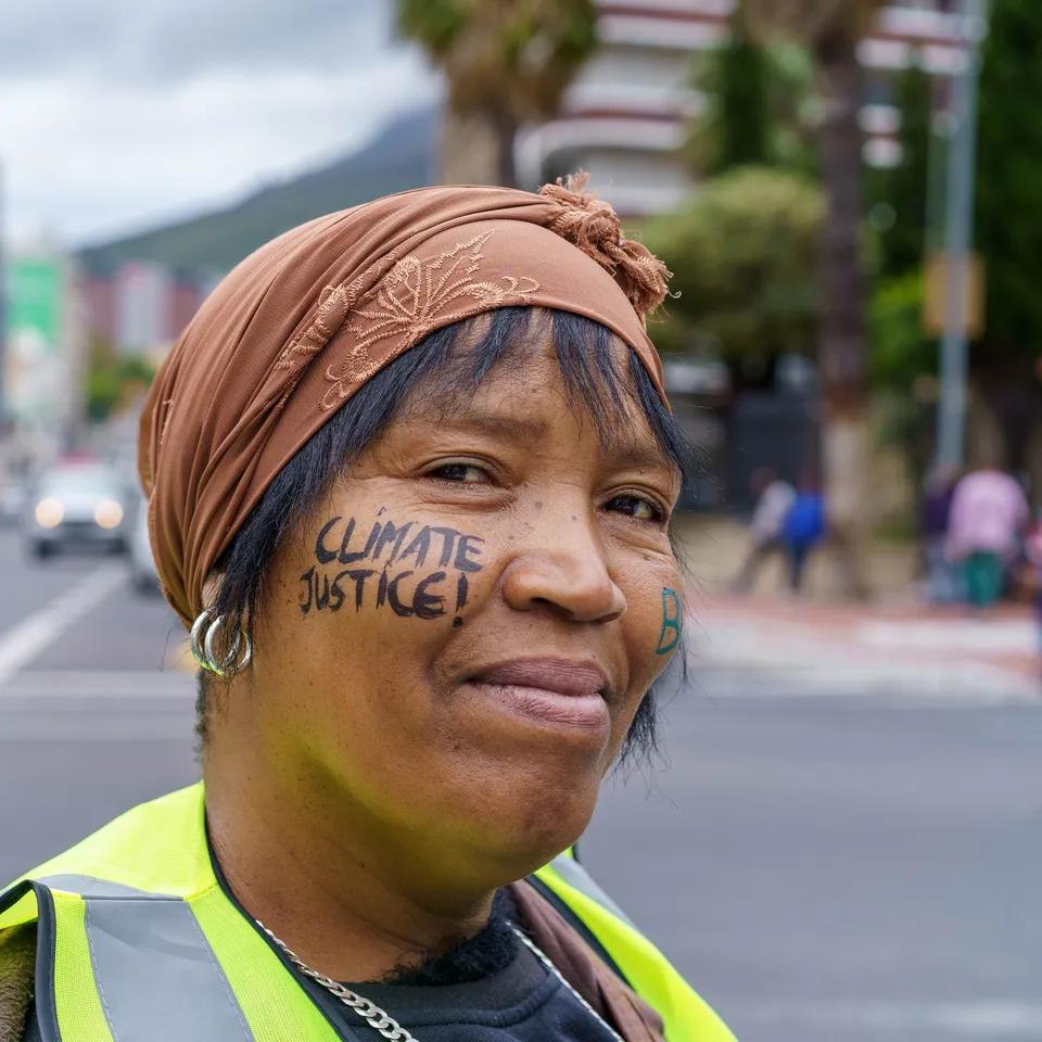 A climate activist wearing a safety vest and orange headwrap stands on an urban street with "CLIMATE JUSTICE!" written on her face, advocating for environmental action in a city setting.