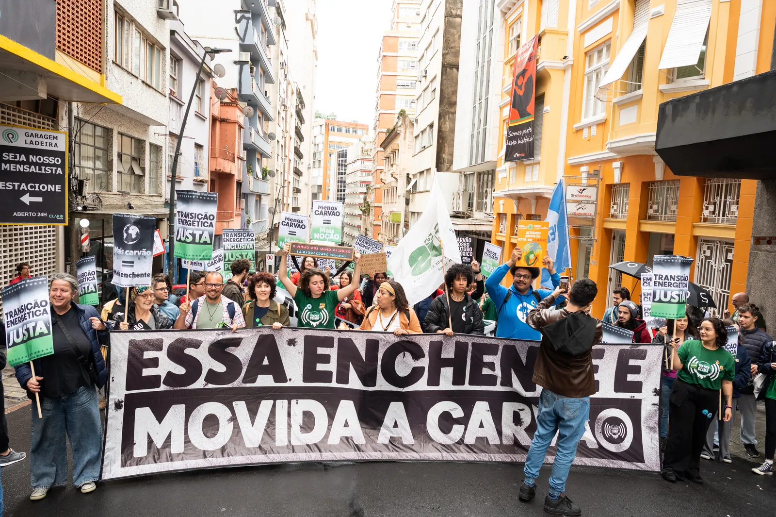 A crowd of protesters marches down a city street holding a large banner with Portuguese text and various signs. Buildings line both sides of the street, with yellow and white facades visible. The demonstration appears organized and peaceful.