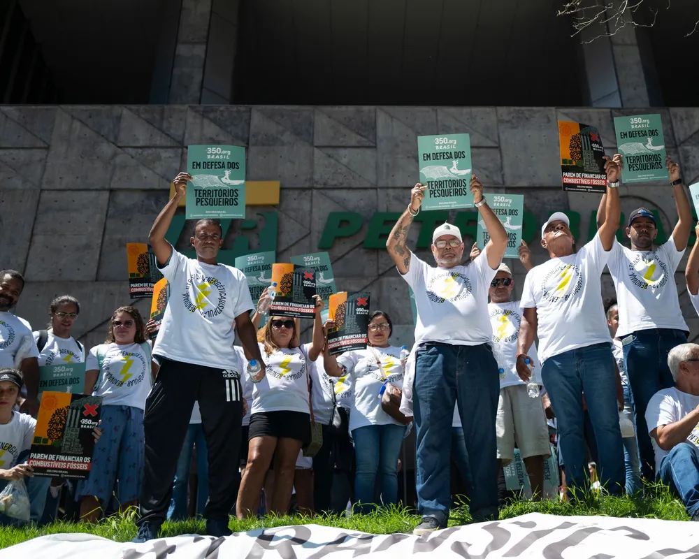 Protesters in white t-shirts with yellow ribbons rally outdoors, holding green signs above their heads. They stand on steps in front of a modern building, demonstrating in an organized group formation.