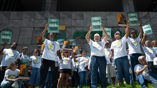 Protesters in white t-shirts with yellow ribbons rally outdoors, holding green signs above their heads. They stand on steps in front of a modern building, demonstrating in an organized group formation.