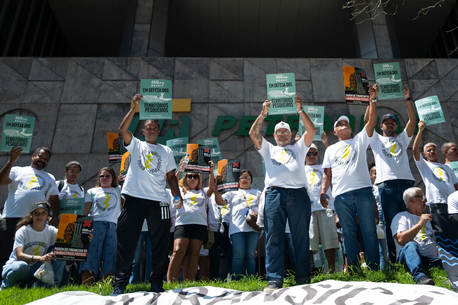 Protesters in white t-shirts with yellow ribbons rally outdoors, holding green signs above their heads. They stand on steps in front of a modern building, demonstrating in an organized group formation.
