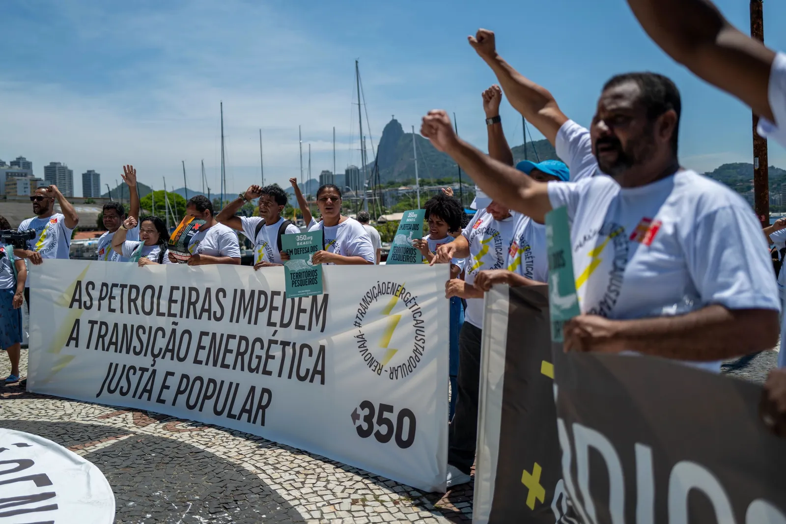Protesters in Rio de Janeiro hold a banner demanding fair energy transition, with iconic Sugarloaf Mountain visible in background. Demonstrators wear white shirts and raise fists in solidarity during the climate action rally.