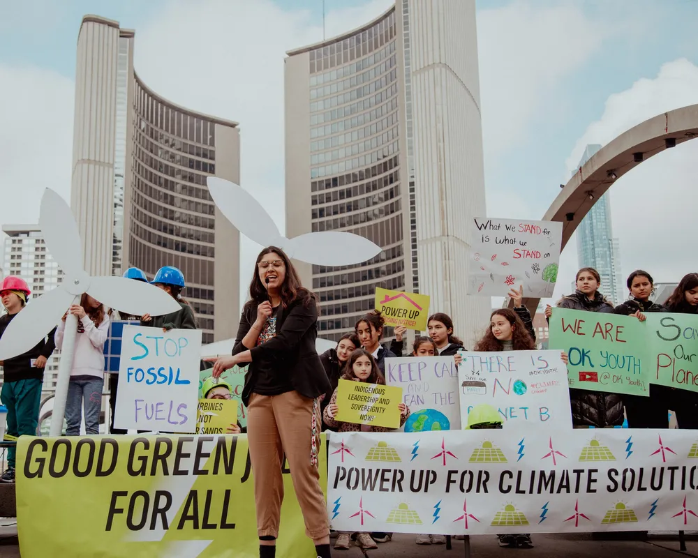 Climate activists rally at Toronto City Hall, holding signs demanding green energy and climate action. A speaker addresses the crowd with iconic curved towers in the background under cloudy skies.