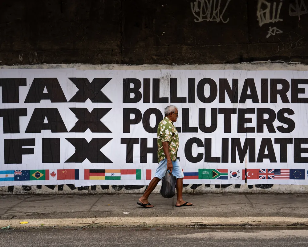 A person walks past a large protest banner reading "TAX BILLIONAIRES TAX POLLUTERS FIX THE CLIMATE" with G20 country flags displayed below. The sign advocates for climate action through taxation of the wealthy.