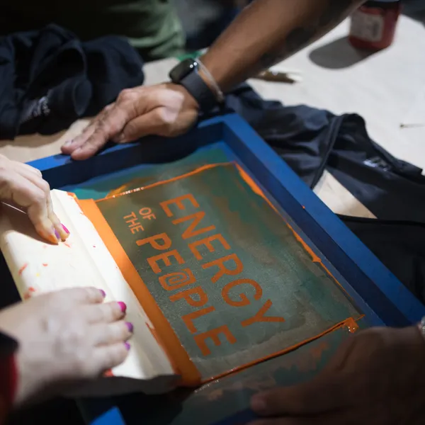 Multiple hands collaborate on screen printing "ENERGY OF THE PEOPLE" text onto fabric.