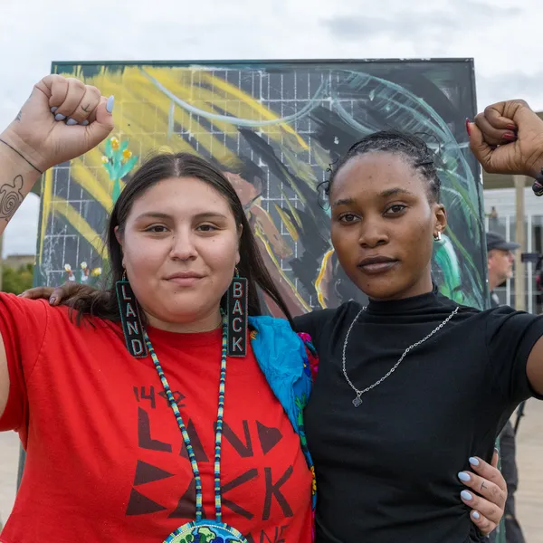 Two women stand together with raised fists symbolizing solidarity. One wears a red "Land Back" shirt, the other in black.