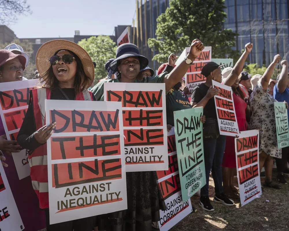 Protesters march outdoors holding "DRAW THE LINE AGAINST INEQUALITY" signs with raised fists, demonstrating for social justice in an urban setting with buildings and trees visible in the background.