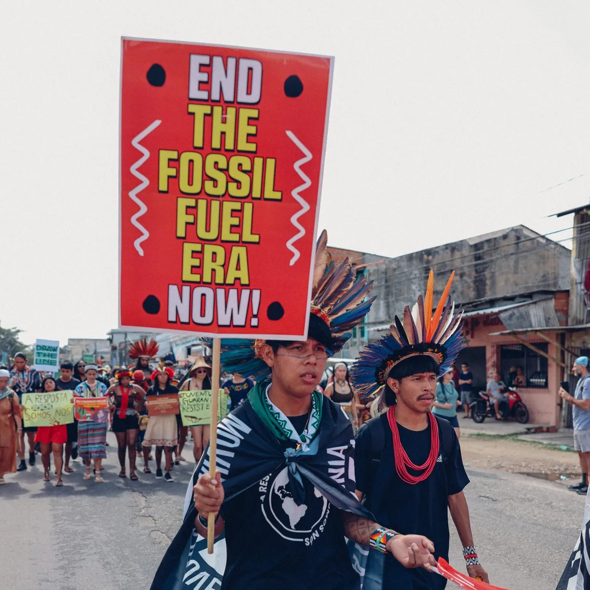 Climate activists march in traditional indigenous dress carrying a bold red and yellow sign demanding an end to the fossil fuel era, leading a street demonstration for environmental justice.