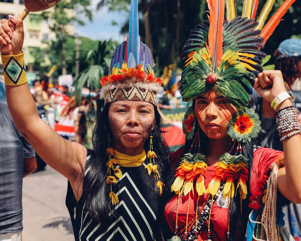Two indigenous women in traditional ceremonial dress with feathered headdresses and colorful costumes participate in a cultural celebration or protest in an urban setting with palm trees and buildings in the background.