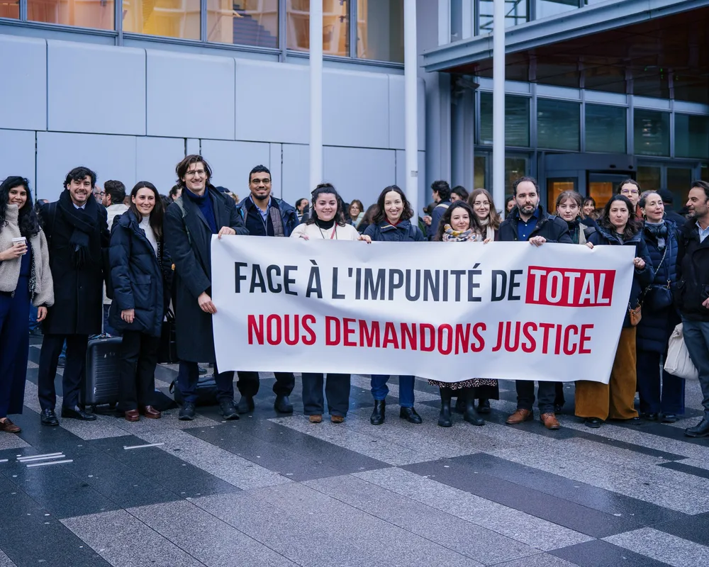 A group of protesters stands outside a modern building holding a French banner that reads "FACE À L'IMPUNITÉ DE TOTAL NOUS DEMANDONS JUSTICE" (Facing Total's impunity, we demand justice), advocating for corporate accountability.