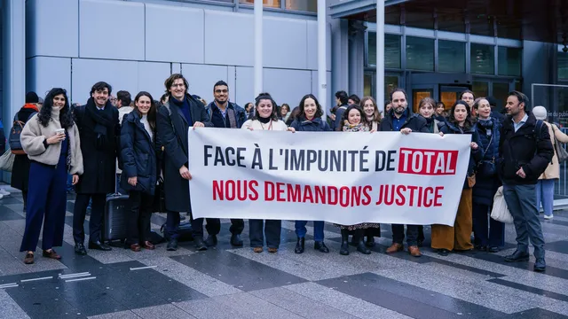 A group of protesters stands outside a modern building holding a French banner that reads "FACE À L'IMPUNITÉ DE TOTAL NOUS DEMANDONS JUSTICE" (Facing Total's impunity, we demand justice), advocating for corporate accountability.