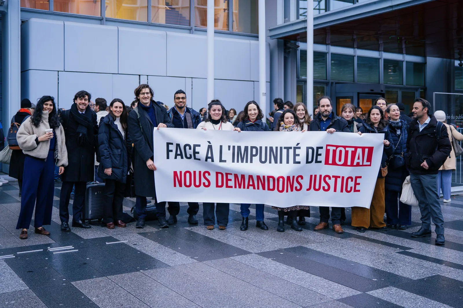 A group of protesters stands outside a modern building holding a French banner that reads "FACE À L'IMPUNITÉ DE TOTAL NOUS DEMANDONS JUSTICE" (Facing Total's impunity, we demand justice), advocating for corporate accountability.