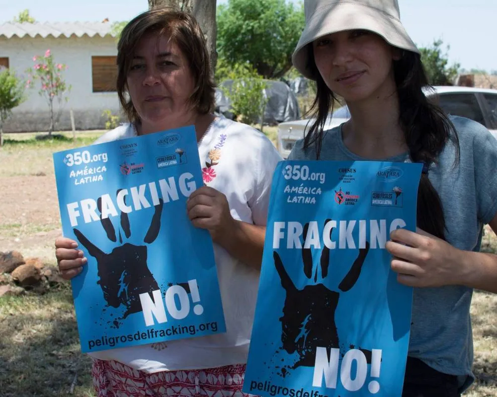 Two women hold blue anti-fracking protest signs reading "FRACKING NO!" with handprint symbols. They stand outdoors in a residential area, participating in an environmental activism campaign by 350.org América Latina.