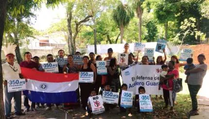 Community group holding protest signs and Paraguayan flag outdoors under trees, advocating for the Chaco Guasu forest with "350.org" placards displayed by participants of various ages.
