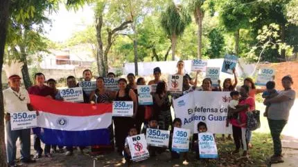 Community group holding protest signs and Paraguayan flag outdoors under trees, advocating for the Chaco Guasu forest with "350.org" placards displayed by participants of various ages.