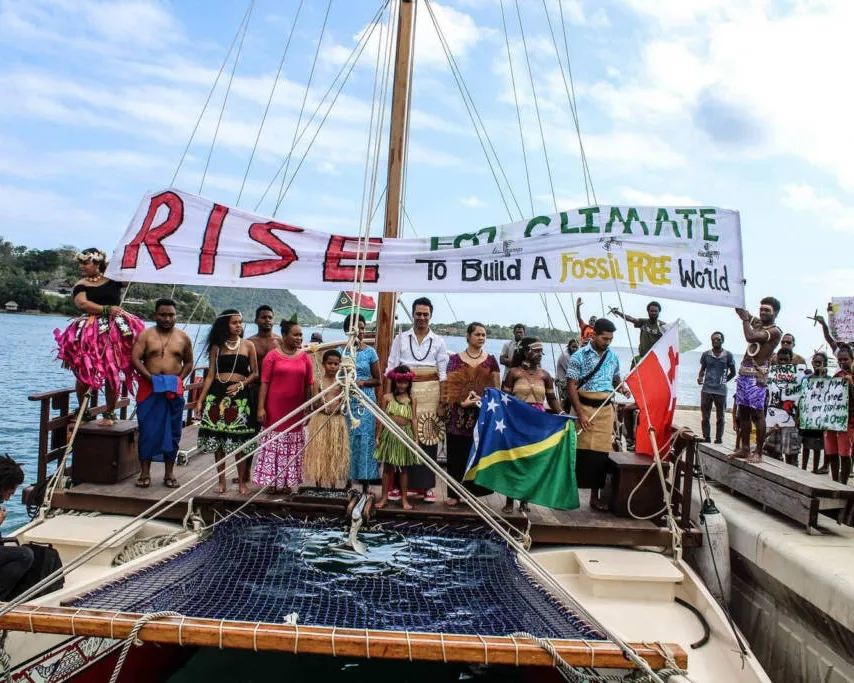 Pacific Islanders gather on a traditional sailing vessel holding banners reading "RISE FOR CLIMATE" and "To Build A Fossil FREE World," protesting climate change impact on island nations.
