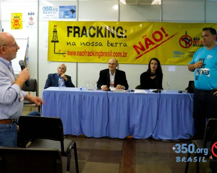 Anti-fracking panel discussion in Brazil with speakers at table beneath yellow "FRACKING: na nossa terra NÃO!" banner. Community members stand and sit while addressing the panel about environmental concerns.