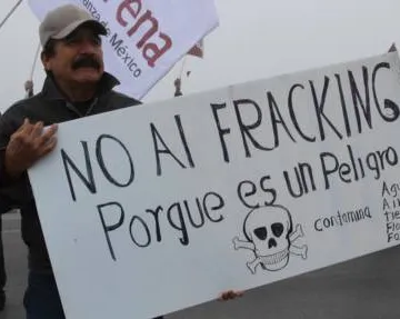 A protester holds a Spanish-language sign opposing fracking, featuring a skull and crossbones symbol with "NO AL FRACKING Porque es un Peligro" (No to fracking because it is a danger) written on it.