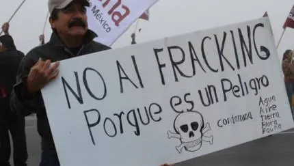 A protester holds a Spanish-language sign opposing fracking, featuring a skull and crossbones symbol with "NO AL FRACKING Porque es un Peligro" (No to fracking because it is a danger) written on it.