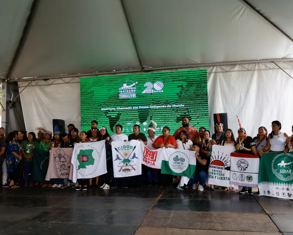 A large group of Indigenous people and activists gather under a white tent, holding organizational banners and flags at what appears to be a community event or assembly focused on Indigenous rights and territorial issues.