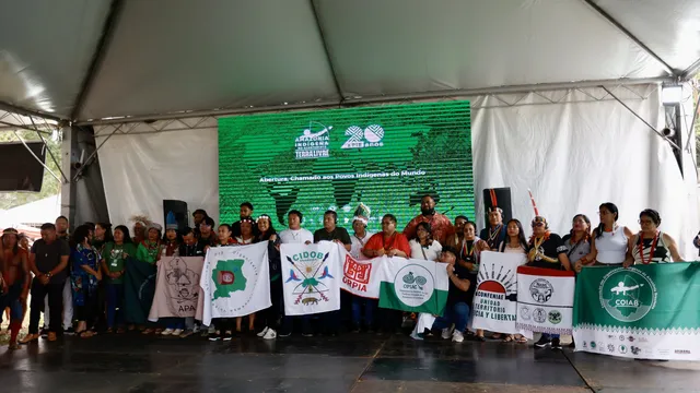 A large group of Indigenous people and activists gather under a white tent, holding organizational banners and flags at what appears to be a community event or assembly focused on Indigenous rights and territorial issues.