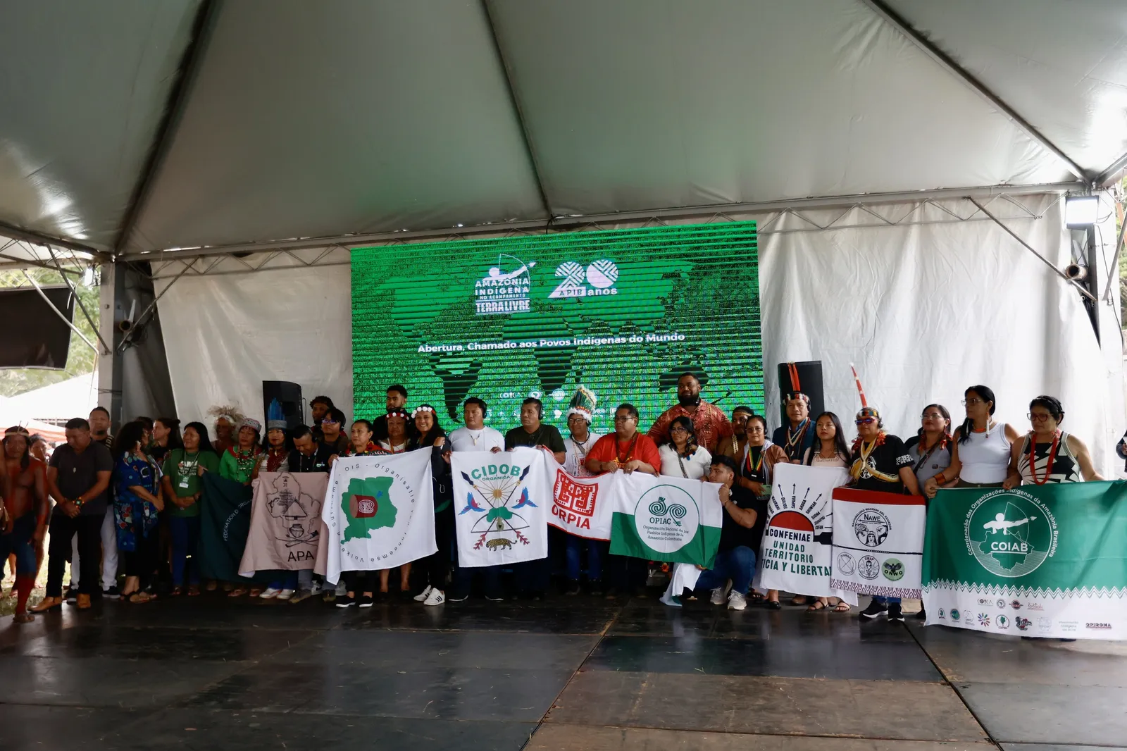 A large group of Indigenous people and activists gather under a white tent, holding organizational banners and flags at what appears to be a community event or assembly focused on Indigenous rights and territorial issues.