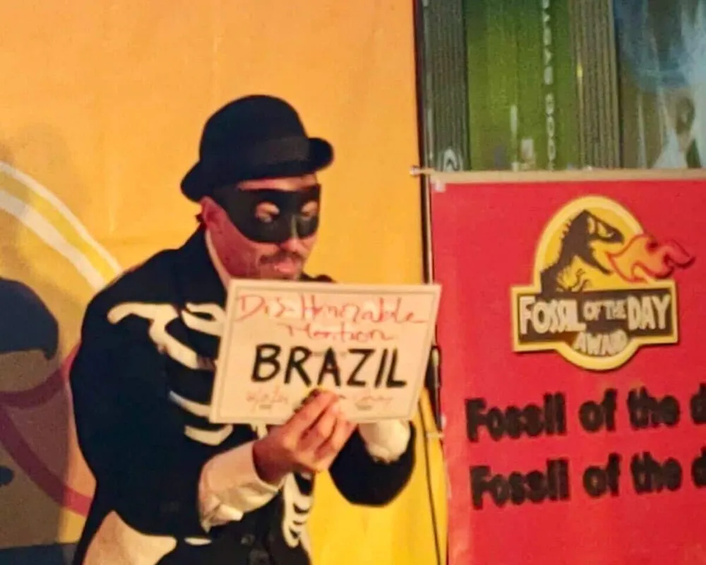 A person in a burglar costume with black mask and striped shirt holds a sign reading "BRAZIL" at what appears to be a themed party or event with colorful backdrop and "Fossil of the Day" signage.