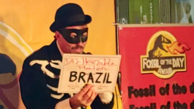 A person in a burglar costume with black mask and striped shirt holds a sign reading "BRAZIL" at what appears to be a themed party or event with colorful backdrop and "Fossil of the Day" signage.