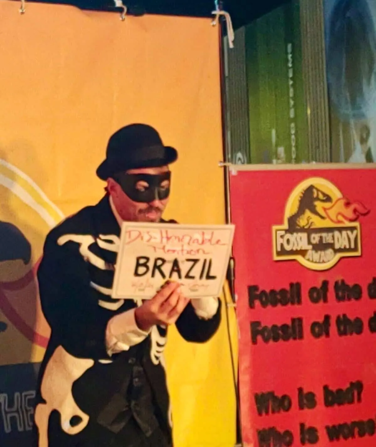 A person in a burglar costume with black mask and striped shirt holds a sign reading "BRAZIL" at what appears to be a themed party or event with colorful backdrop and "Fossil of the Day" signage.