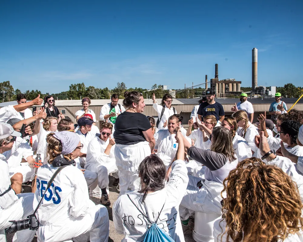 A large group of climate activists protests and puts their fists in the air in front of a coal plant in New Hampshire.
