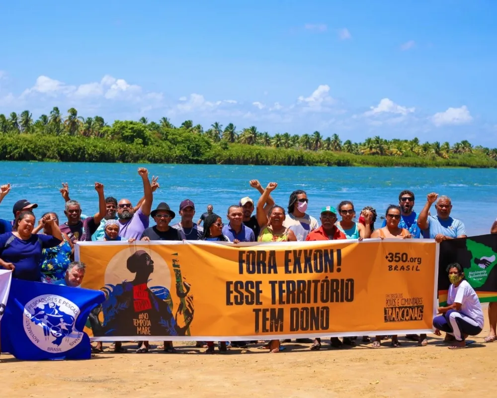Community members gather on a beach to protest, holding a large orange banner reading "FORA EXXON! ESSE TERRITORIO TEM DONO" against oil drilling, with a river and palm trees in the background under blue skies.