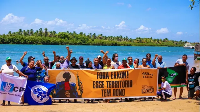 Community members gather on a beach to protest, holding a large orange banner reading "FORA EXXON! ESSE TERRITORIO TEM DONO" against oil drilling, with a river and palm trees in the background under blue skies.