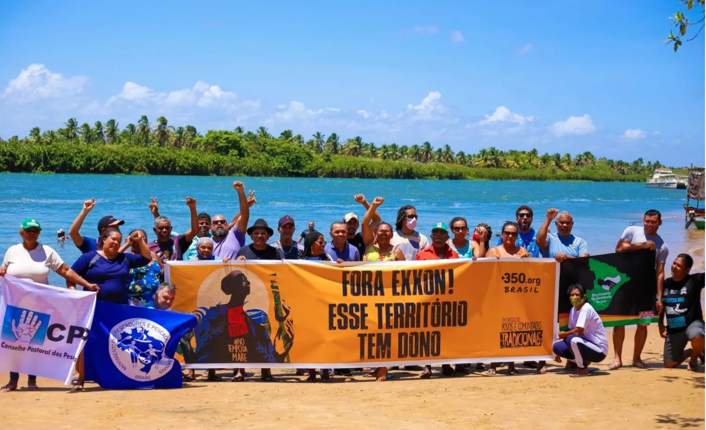 Community members gather on a beach to protest, holding a large orange banner reading "FORA EXXON! ESSE TERRITORIO TEM DONO" against oil drilling, with a river and palm trees in the background under blue skies.