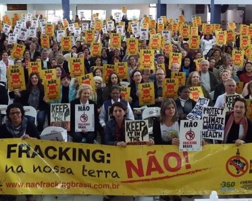 Large crowd of protesters holding yellow and white signs reading "NÃO" (No) and anti-fracking messages, gathered behind a yellow banner opposing fracking in Brazil. Democratic demonstration for environmental protection.