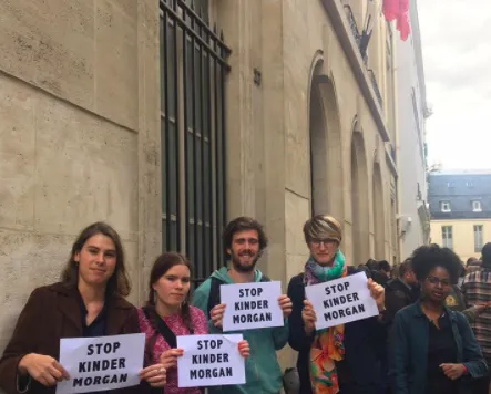 A group of five protesters stand outside a government building in Paris, holding "STOP KINDER MORGAN" signs. They appear to be demonstrating against the Kinder Morgan pipeline project, with French and EU flags visible in the background.