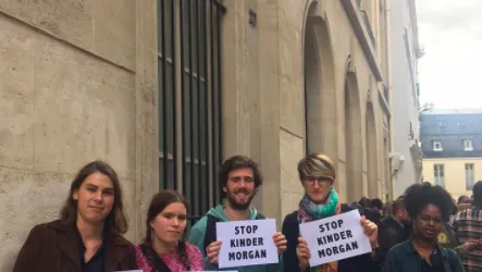 A group of five protesters stand outside a government building in Paris, holding "STOP KINDER MORGAN" signs. They appear to be demonstrating against the Kinder Morgan pipeline project, with French and EU flags visible in the background.