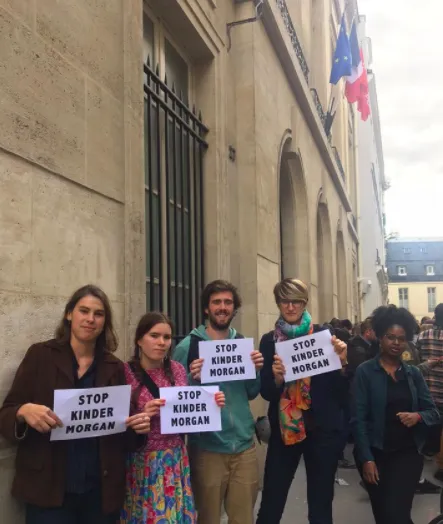 A group of five protesters stand outside a government building in Paris, holding "STOP KINDER MORGAN" signs. They appear to be demonstrating against the Kinder Morgan pipeline project, with French and EU flags visible in the background.