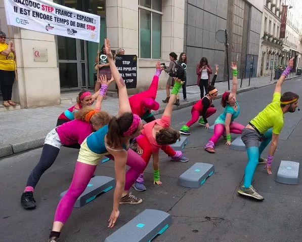 Women in colorful 80s-style workout attire perform aerobics with step platforms on a city street during a protest event. Banners about Gazoduc STEP France-Espagne are visible in the background.
