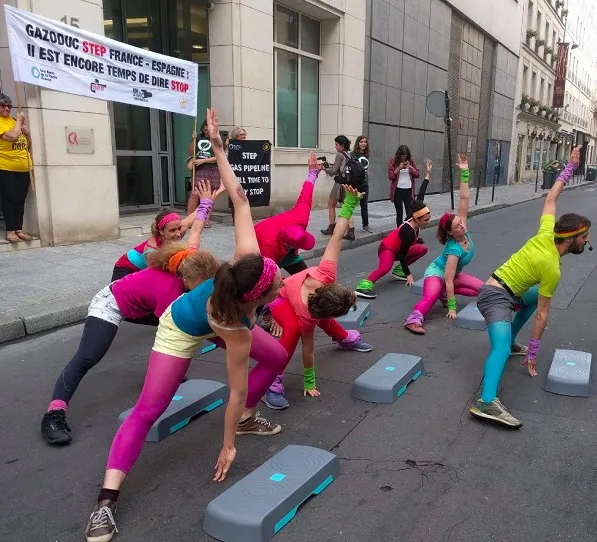 Women in colorful 80s-style workout attire perform aerobics with step platforms on a city street during a protest event. Banners about Gazoduc STEP France-Espagne are visible in the background.