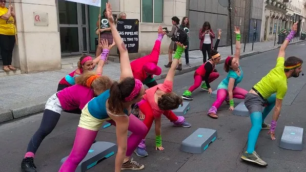 Women in colorful 80s-style workout attire perform aerobics with step platforms on a city street during a protest event. Banners about Gazoduc STEP France-Espagne are visible in the background.