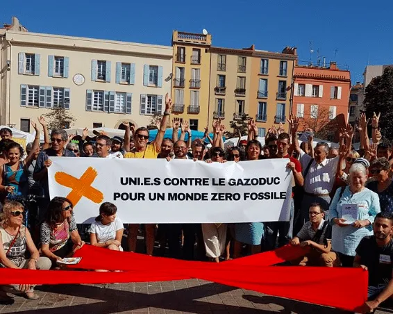 A large group of environmental protesters gathered in a town square holding a banner in French that reads "United against gas pipelines for a zero fossil fuel world" with arms raised in solidarity.