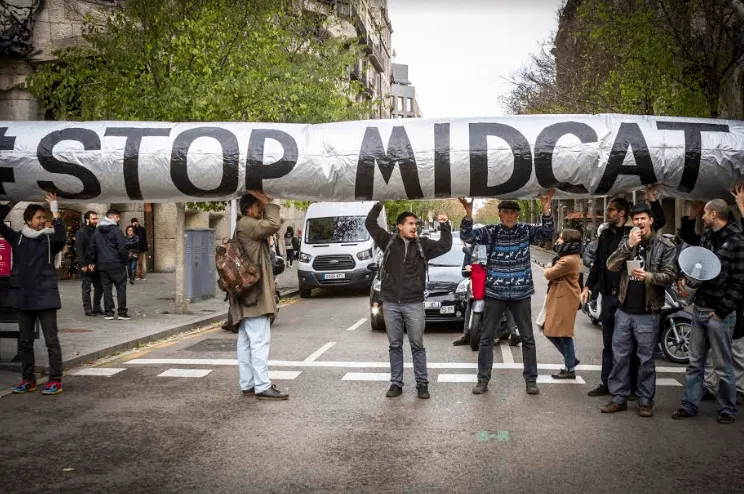 Protesters hold a large "STOP MIDCAT" banner across a street during a demonstration. People gather on both sides as participants display their opposition to the MIDCAT pipeline project in an urban setting.