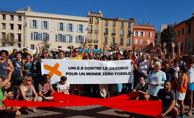 A large group of environmental protesters gathered in a town square holding a banner in French that reads "United against gas pipelines for a zero fossil fuel world" with arms raised in solidarity.