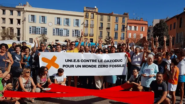 A large group of environmental protesters gathered in a town square holding a banner in French that reads "United against gas pipelines for a zero fossil fuel world" with arms raised in solidarity.