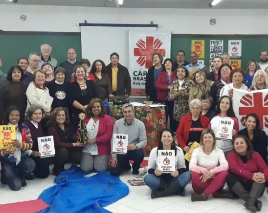 Large group photo of diverse people gathered in a meeting room with Caritas organization banners and posters displayed. Participants are arranged in rows, some seated on floor, others standing, in casual attire.