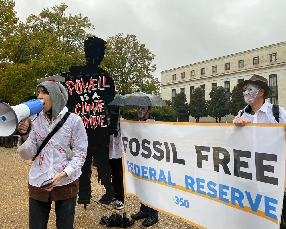 In the rain outside the DC Federal Reserve building, A cutout of a person that says "Powell is a Climate Zombie," next to two people holding a banner reading "Fossil Free Federal Reserve" behind a person speaking into a megaphone wearing a Zombie costume.