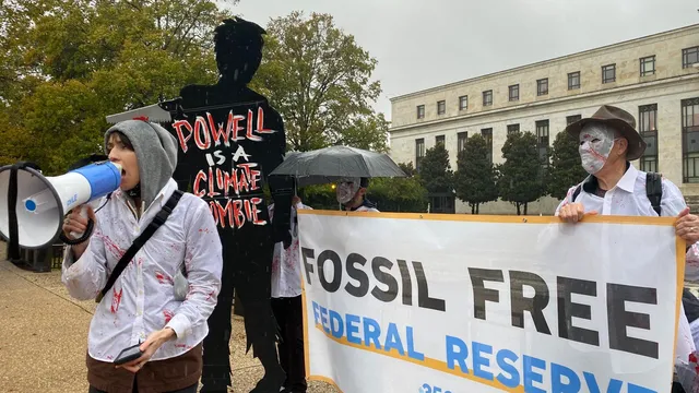 In the rain outside the DC Federal Reserve building, A cutout of a person that says "Powell is a Climate Zombie," next to two people holding a banner reading "Fossil Free Federal Reserve" behind a person speaking into a megaphone wearing a Zombie costume.