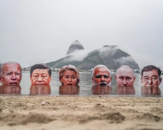 Six life-sized cutouts of world leaders' heads partially submerged in water at a beach, with misty mountains in the background, creating a striking political art installation.
