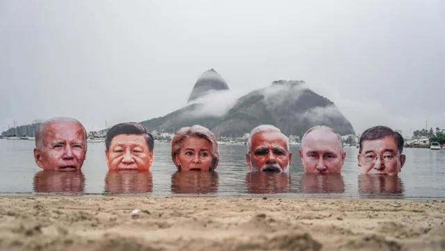 Six life-sized cutouts of world leaders' heads partially submerged in water at a beach, with misty mountains in the background, creating a striking political art installation.